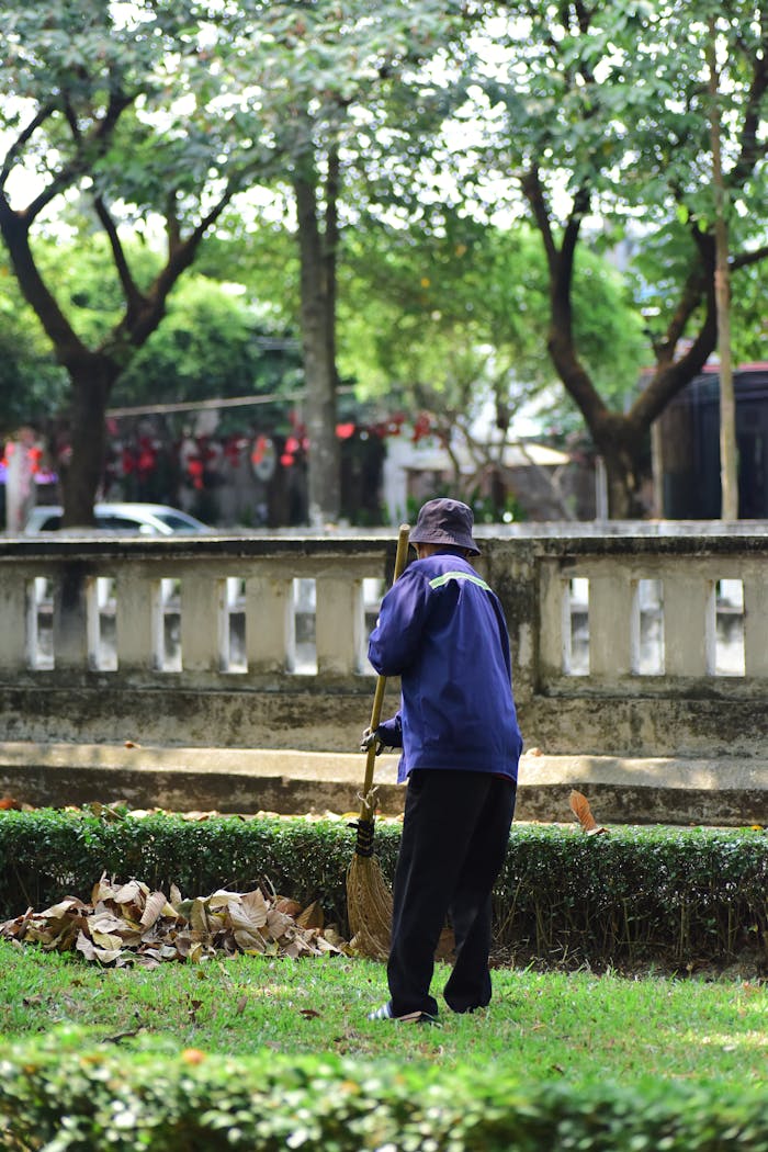 A gardener tidying up leaves in a serene park setting with trees and greenery.